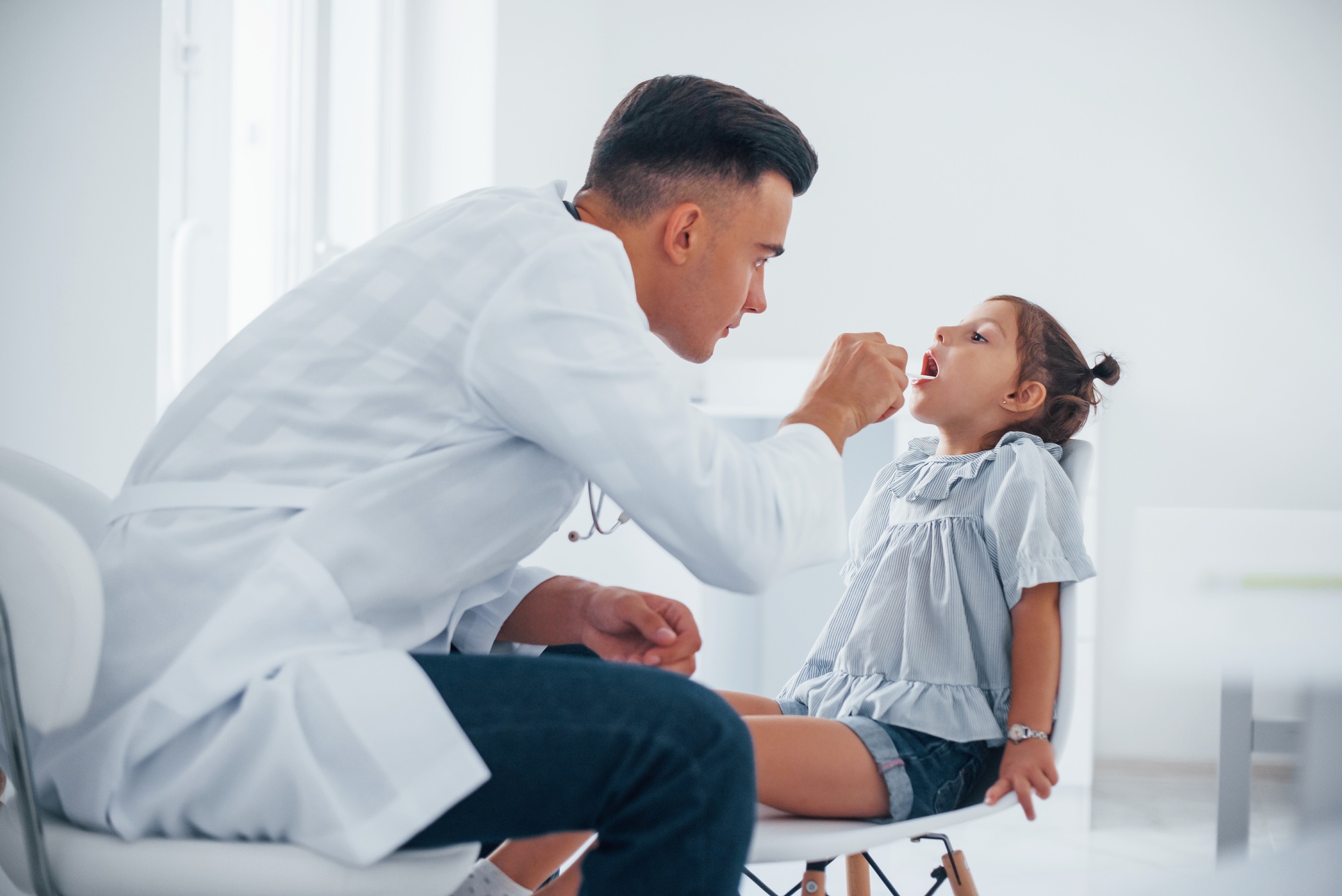 Uses tonsil to check throat. Young pediatrician works with little female visitor in the clinic